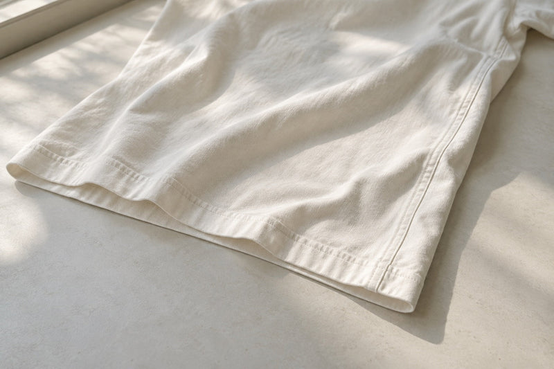 AI-generated editorial illustration of an ivory T-shirt side seam and twin-needle hem in natural light; not a product photograph or wash test.